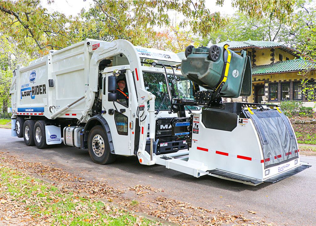 A white Heil Low Rider automated refuse truck lifts a green residential trash bin using its side loading arm. The truck is on a tree lined suburban street with a house in the background.