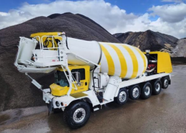 A large yellow and white Terex concrete mixer truck is parked in an industrial area with gravel piles and mountains behind it. The truck has an extended rotating drum and multiple rear axles for heavy duty operation.