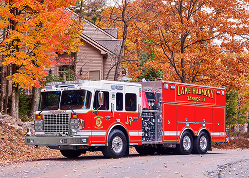 A red Lake Harmony Tanker 17 fire truck is parked on a residential street surrounded by bright autumn foliage. The truck features a large water tank, hose controls, and a white cab with chrome details.